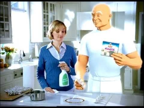 a woman holding a bottle of cleaning agent stands in a kitchen next to Mr. Clean as he cleans the stove
