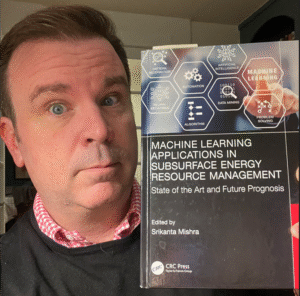 Photo of Zack Warren holding up a book titled “Machine Learning Applications in Subsurface Energy Resource Management: State of the Art and Future Prognosis,” edited by Srikanta Mishra (CRC Press). He looks wide-eyed at the camera while presenting the book, which features icons related to artificial intelligence, neural networks, algorithms, and data mining on the cover.